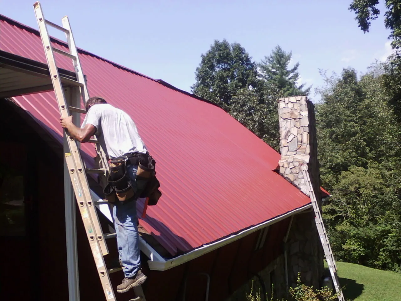 Roofer on a ladder beside a red metal roof and stone chimney — Soffit & Fascia Repair by Roofing Brothers in Thomson